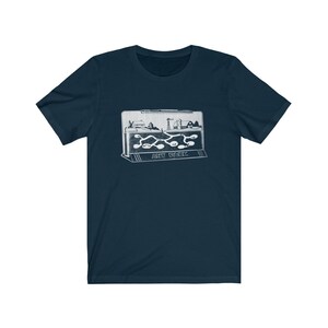 May include: A navy blue t-shirt with a white graphic of an ant farm. The graphic shows a small plastic ant farm with a miniature town inside and ants crawling through tunnels. The text "ANT FARM" is printed below the graphic.