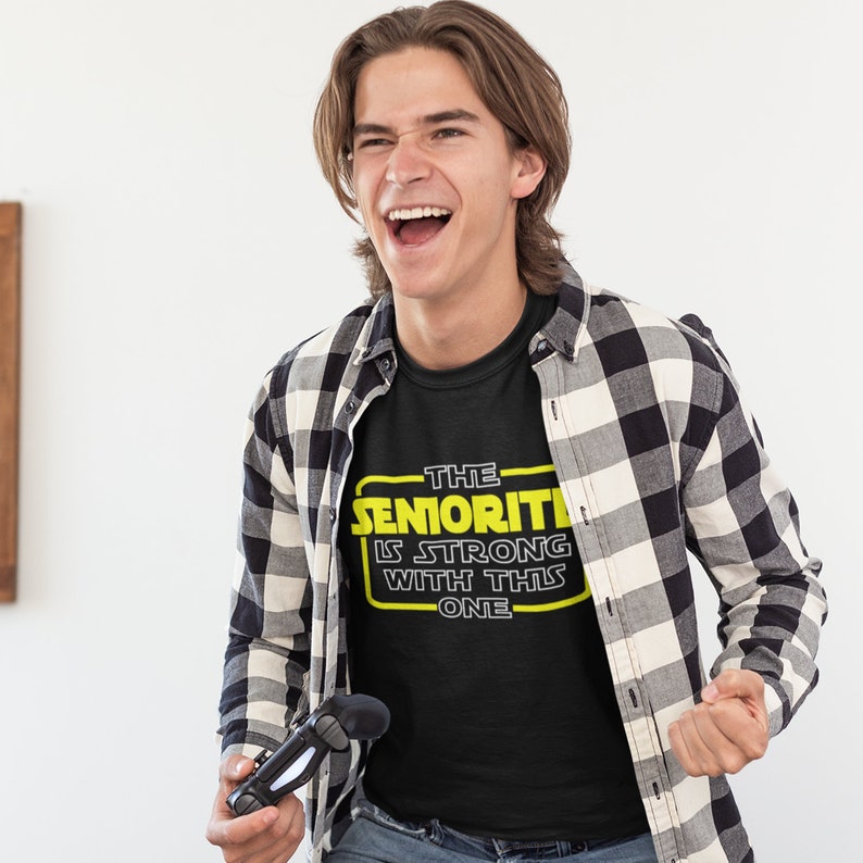 May include: A person wearing a black t-shirt with a yellow graphic that says "The Seniority Is Strong With This One". The person is holding a black video game controller and smiling.