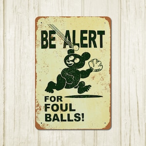 May include: A vintage-style metal sign with a distressed yellow background and green text. The sign features a cartoon bear wearing a baseball glove and running to catch a ball. The text on the sign reads "BE ALERT FOR FOUL BALLS!"