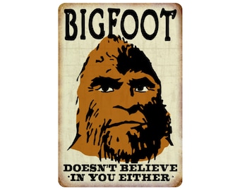 May include: A rectangular metal sign with a distressed finish features a brown Bigfoot face and the text "BIGFOOT" above and "DOESN'T BELIEVE IN YOU EITHER" below. The sign has a vintage, rustic aesthetic.