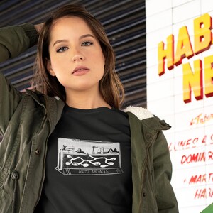 May include: A woman wearing a black t-shirt with a white graphic of an ant farm. The graphic says "Ant Farm". She is wearing a green jacket and has her hand in her hair.