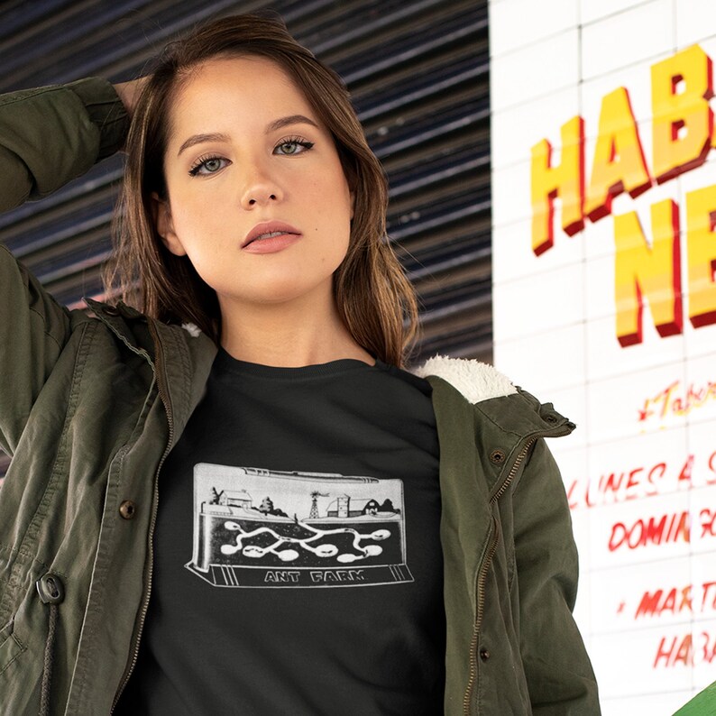 May include: A woman wearing a black t-shirt with a white graphic of an ant farm. The graphic says "Ant Farm". She is wearing a green jacket and has her hand in her hair.