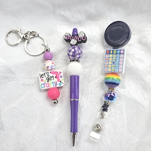Puede incluir: Colección de accesorios con cuentas, que incluye un llavero con un dije "Let's Get Crafty", un bolígrafo morado y un portatarjetas retráctil con un diseño de cartón de bingo. Las cuentas son de color morado, rosa, blanco y arcoíris.