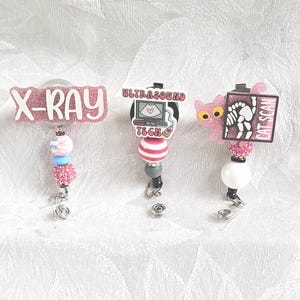 May include: Three medical-themed badge reels. One reads "X-RAY" in pink glitter, another says "ULTRASOUND TECH", and the last says "CAT SCAN". Each has a beaded design with a retractable cord and a metal clip.