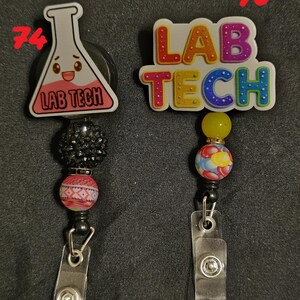 Może przedstawiać: Dwa chowane uchwyty na identyfikatory. Jeden przedstawia kresk&oacute;wkową kolbę z napisem "LAB TECH", drugi ma "LAB TECH" w kolorowych literach. Oba mają ozdobne koraliki i przezroczysty plastikowy klips.