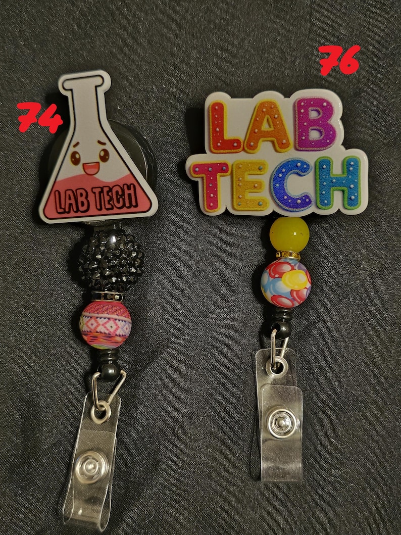 Może przedstawiać: Dwa chowane uchwyty na identyfikatory. Jeden przedstawia kresk&oacute;wkową kolbę z napisem "LAB TECH", drugi ma "LAB TECH" w kolorowych literach. Oba mają ozdobne koraliki i przezroczysty plastikowy klips.