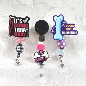 May include: Three medical-themed badge reels with humorous phrases. One reel says "IT'S GOING TIBIA OKAY" with a pink and black design. Another says "Oh Snap!" and the third says "I FOUND THIS HUMERUS".