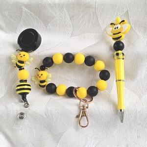 May include: A set of bee-themed accessories, including a badge reel, bracelet, and pen. The badge reel and pen feature yellow and black bee designs. The bracelet has yellow and black beads. The pen has a yellow and black plaid pattern.