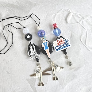 May include: Three retractable badge holders with airplane charms. One has a black and white graphic of a person with luggage and the text "Flight Attendant". Another has a woman in a blue uniform. The third says "Let's Cruise".