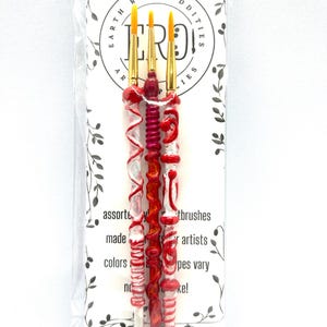 May include: A set of three artist paintbrushes with red and white swirled handles and yellow bristles. The brushes are packaged in a clear plastic sleeve with a white card that reads "EARTH RARE ODDITIES" and "assorted paintbrushes made for artists."