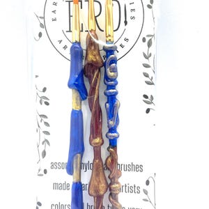 May include: Three paint brushes with blue, brown, and gold handles. The brushes are packaged in a clear plastic bag with a white card that reads "Earth Rare Oddities" and "ERDI".