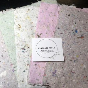 May include: A stack of handmade paper sheets in various pastel colors, speckled with recycled materials and pressed flowers. A white card with the text "HANDMADE PAPER" is on top.
