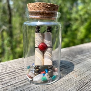 May include: A clear glass jar with a cork stopper, containing two rolled scrolls, a red bead, and colorful stones. The scrolls are beige with dark accents. The jar sits on a wooden surface with a blurred green background.