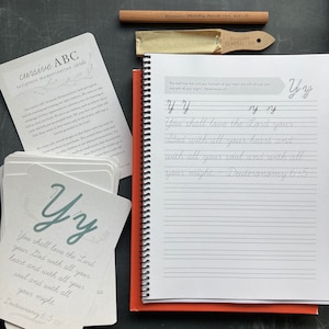 May include: A set of cursive alphabet scripture memorization cards with a green and white floral design. The cards feature the text "You shall love the Lord your God with all your heart and with all your soul and with all your might. - Deuteronomy 6:5".  The cards are laid out on a red notebook with a white page open to the same scripture written in cursive.