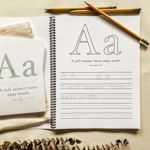 May include: An open spiral-bound notebook and flashcards for learning the letter "A". The notebook features the letter "A" in uppercase and lowercase, with lines for practice. The text "A soft answer turns away wrath" is also included. Pencils are placed on the notebook.