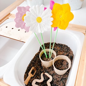 May include: A white, pink and yellow felt flower arrangement in a white bowl filled with brown beans. A small wooden rake and a brown felt worm are also in the bowl.