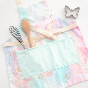 May include: A white apron with a large pocket and a watercolour design of pink, blue, and green clouds with gold stars and moons. The apron has a cream-coloured tie and is shown with a wooden spoon and a whisk.