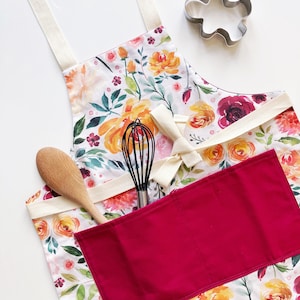 May include: A floral print apron with a red pocket, a wooden spoon, and a whisk. The apron features a watercolour design with orange, yellow, and burgundy flowers. A metal cookie cutter is also visible.