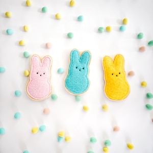 Peut inclure: Trois formes de lapins de Pâques en feutre rose, bleu et jaune. Les lapins sont disposés en rangée sur un fond blanc avec des œufs de bonbons de couleur pastel éparpillés.
