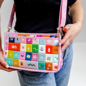 Puede incluir: Un bolso bandolera acolchado rectangular con una cuadrícula de cuadrados de tela coloridos con varios iconos blancos pequeños, incluyendo un ancla, un búho, estrellas y frutas. El bolso tiene una correa y un borde de color rosa claro.
