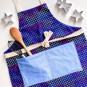 May include: A blue and rainbow heart patterned apron with a large blue pocket and white ties. The apron has a wooden spoon and a whisk in the pocket.