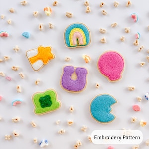 Può includere: Modello di ricamo per toppe di feltro con un arcobaleno, una stella, un ferro di cavallo, un trifoglio, una nuvola e una luna crescente. Le toppe sono disposte su uno sfondo bianco con pezzi di cereali sparsi.