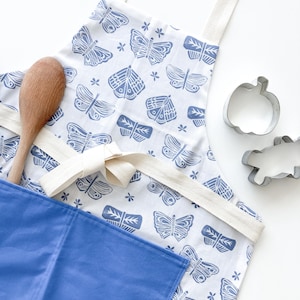 May include: A blue and white apron with a butterfly print. The apron has a large pocket on the front and a white tie.
