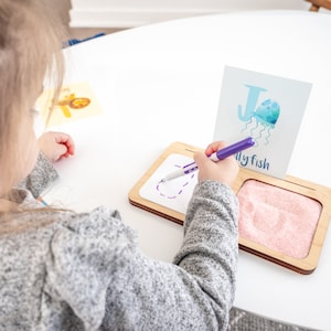 May include: A child traces the letter J on a white dry-erase board with a purple marker. The letter J is part of a learning card with a blue and white watercolour illustration of a jellyfish and the word "jellyfish". The board has a separate section filled with pink sand.