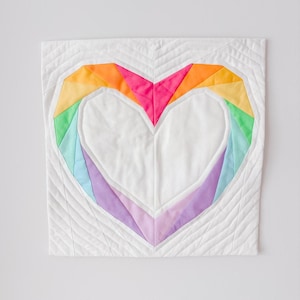 May include: A quilted wall hanging featuring a large heart design. The heart is outlined with a rainbow of colors, including pink, orange, yellow, green, blue, and purple. The background is white with a quilted pattern.