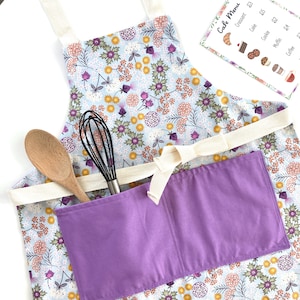 May include: A light blue apron with a floral print and a purple pocket. The apron has a white strap and a large pocket on the front. A wooden spoon and a whisk are in the pocket.