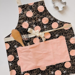 May include: A black and gold apron with a floral pattern and orange citrus fruit design. The apron has a large pocket on the front and is tied with a white ribbon.