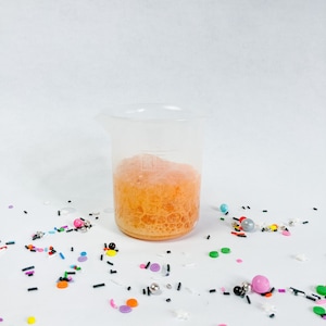 May include: A clear glass beaker filled with a foamy, orange liquid. The beaker is sitting on a white surface surrounded by colourful sprinkles.