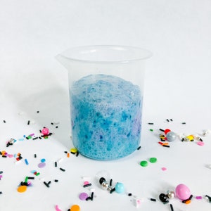 May include: A clear plastic beaker filled with blue and purple foamy liquid. The beaker is sitting on a white surface with colourful sprinkles scattered around it.