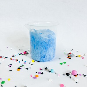 May include: A clear plastic beaker filled with light blue foamy liquid. The beaker is surrounded by colourful sprinkles and small beads.