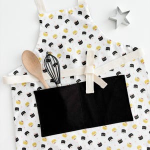 May include: White cotton apron with a black and yellow sushi print. The apron has a large black pocket with a white trim.