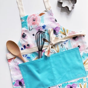 May include: A blue and white floral print apron with a large turquoise pocket. The apron has a white tie and a whisk is in the pocket.