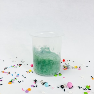 May include: A clear plastic measuring cup with a green powder inside. The cup has markings up to 200 millilitres. The cup is surrounded by colourful sprinkles.