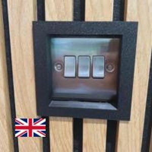 May include: A black light switch plate with three silver toggle switches. The plate is set into a wooden panel with vertical grooves. A Union Jack flag is visible in the lower left corner.