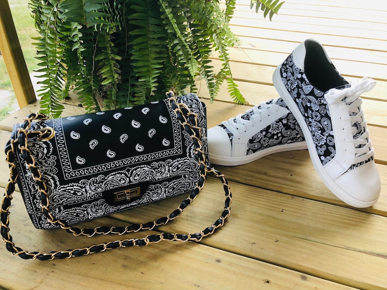 Pu&ograve; includere: Una borsa a tracolla nera e bianca con stampa bandana, con una tracolla a catena dorata e un paio di scarpe da ginnastica bianche con stampa bandana nera e bianca.