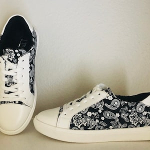 Pu&ograve; includere: Un paio di scarpe da ginnastica in tela bianca con stampa paisley nera. Le scarpe hanno lacci bianchi e una suola in gomma bianca.