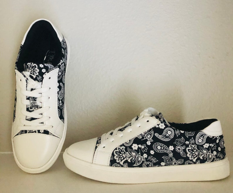 Pu&ograve; includere: Un paio di scarpe da ginnastica in tela bianca con stampa paisley nera. Le scarpe hanno lacci bianchi e una suola in gomma bianca.
