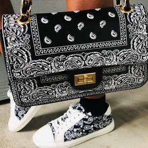 Pu&ograve; includere: Una borsa a mano nera e bianca con stampa bandana, con una tracolla a catena dorata e una chiusura dorata. La borsa &egrave; indossata da una persona che indossa scarpe da ginnastica bianche con stampa bandana nera e bianca.