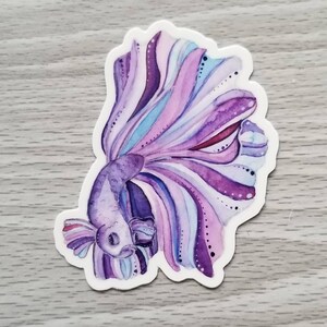 Peut inclure: Un autocollant en aquarelle violet et bleu d'un poisson combattant avec une longue queue flottante.