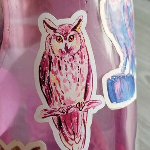 May include: A pink and white watercolor owl sticker with a detailed design. The owl is perched on a branch with its wings spread out.