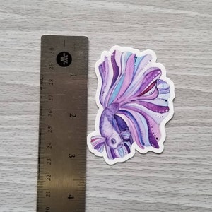 Peut inclure: Un autocollant en aquarelle violet et bleu d'un poisson combattant avec une longue queue flottante.