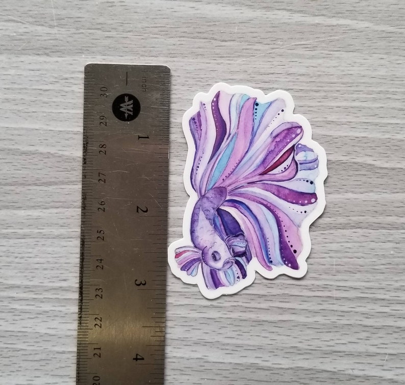 Peut inclure: Un autocollant en aquarelle violet et bleu d'un poisson combattant avec une longue queue flottante.