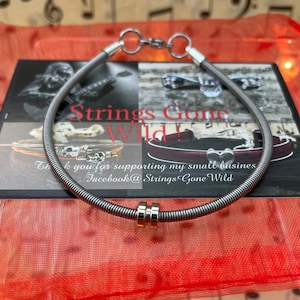 May include: A silver-coloured necklace crafted from coiled metal, with a clasp and two gold-coloured beads. The necklace is displayed on a card with the text "Strings Gone Wild!" and a Facebook address, against a red backdrop.