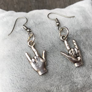 May include: A pair of silver earrings with a rock and roll hand gesture charm. The earrings are on a white fuzzy surface.