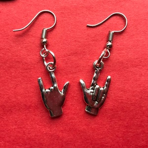 May include: A pair of silver earrings featuring a hand making the 'I love you' sign. The earrings are on a red background.