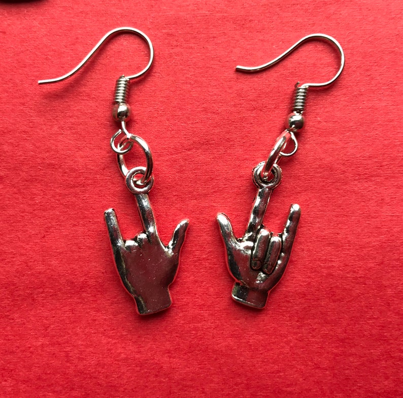 May include: A pair of silver earrings featuring a hand making the 'I love you' sign. The earrings are on a red background.